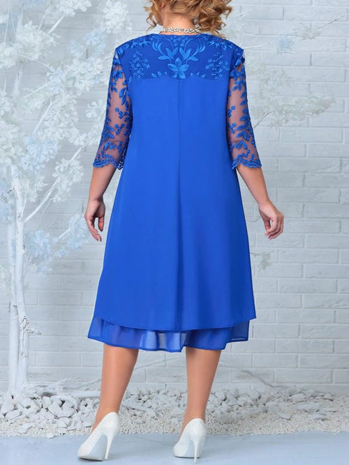 Woman wearing a blue dress with lace sleeves against a white brick wall.