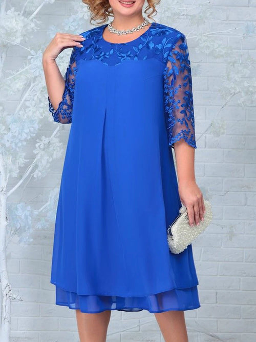 Woman wearing a blue dress with lace sleeves against a white brick wall.